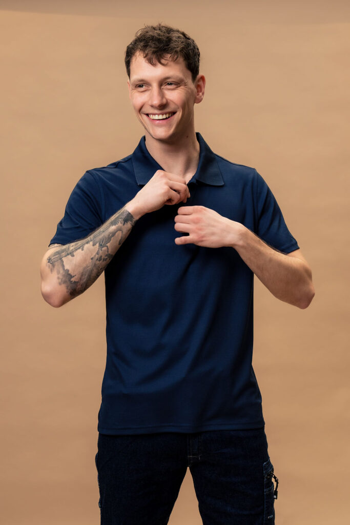 Man draagt Polo Lennox donkerblauw. 100% circulaire bedrijfskleding. Hij pakt de knoop bij zijn kraag met twee handen en kijkt lachend links van de camera. Hij heeft een tatoeage op zijn rechterarm.